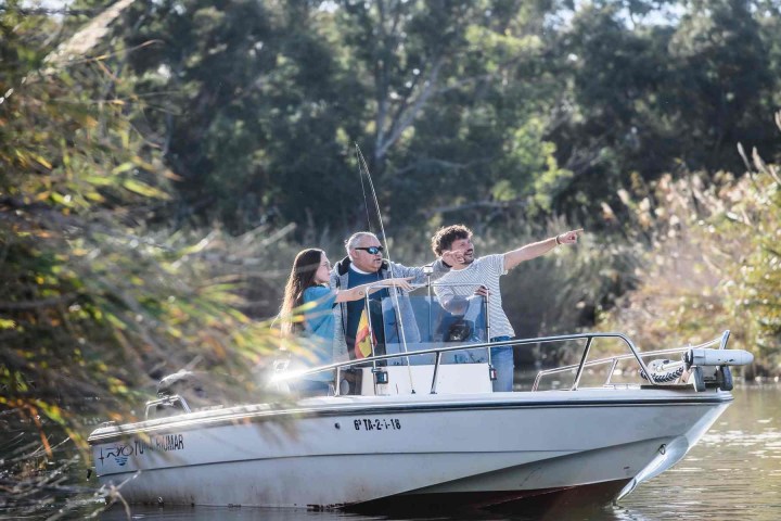 Three people on a small boat in a river, pointing and looking at something in the distance.