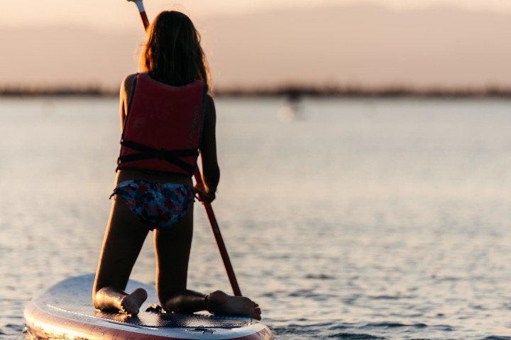 Person kneeling on a paddleboard in the water at sunset.