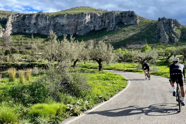 Two cyclists on a winding road with rocky hills and greenery in the background.